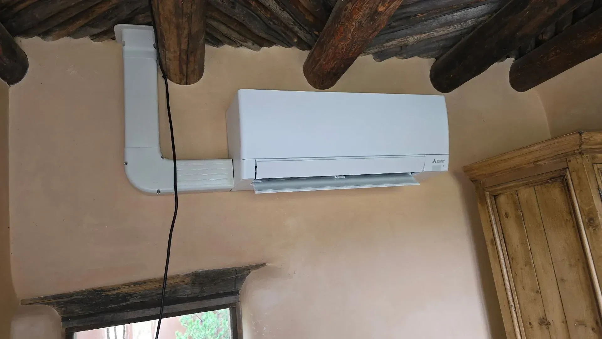 White air conditioning unit mounted on a tan wall, connected by a duct to the ceiling.