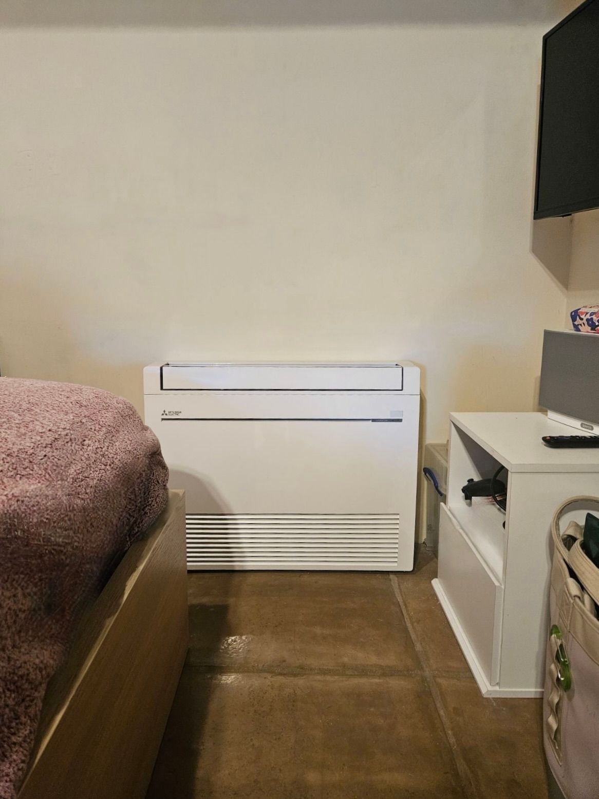 White air conditioning unit against a beige wall in a bedroom with a bed and nightstand.