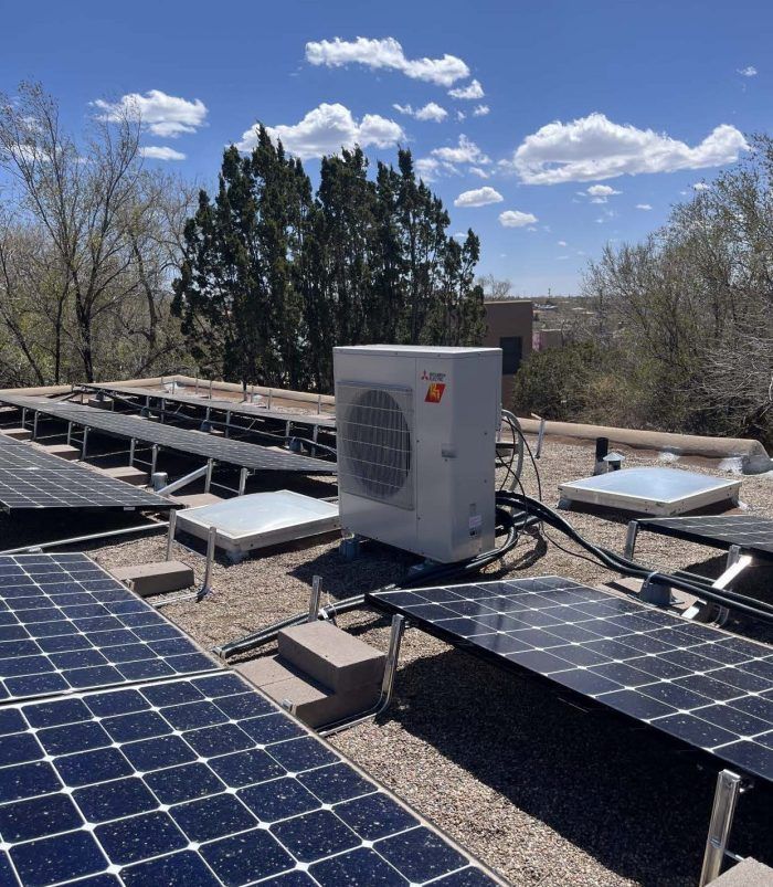 Rooftop solar panels surround an air conditioning unit. Clear blue sky with puffy clouds.