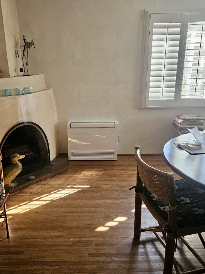 A living room with fireplace, air conditioner, window with shutters, wood floor, and chair.