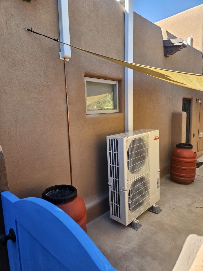 An air conditioning unit next to a brown stucco building with a barrel and blue gate.