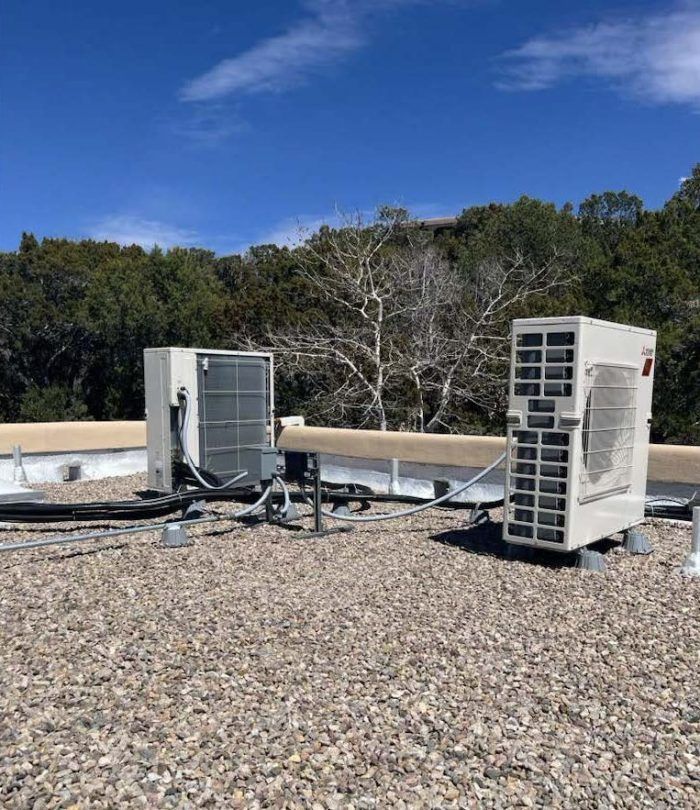 Two white HVAC units on a gravel roof, with trees and blue sky in the background.