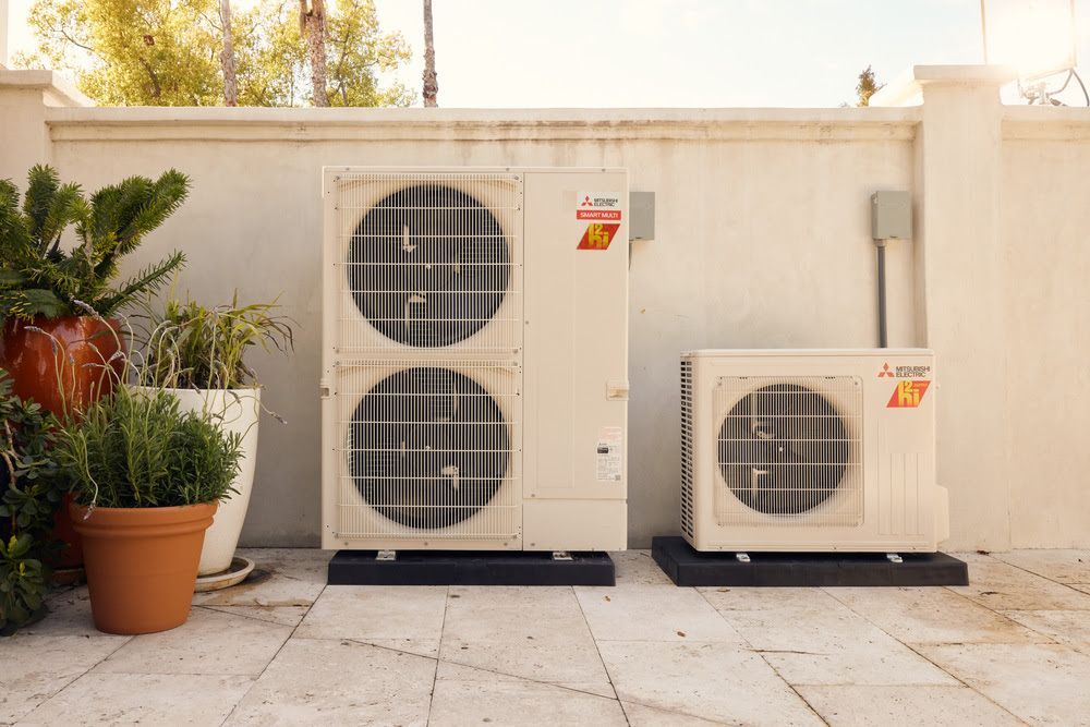 Two white air conditioning units on black pads against a white wall with plants in pots.