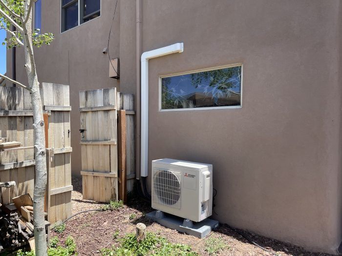 An air conditioning unit and white drain pipe on a tan building's exterior with a window.