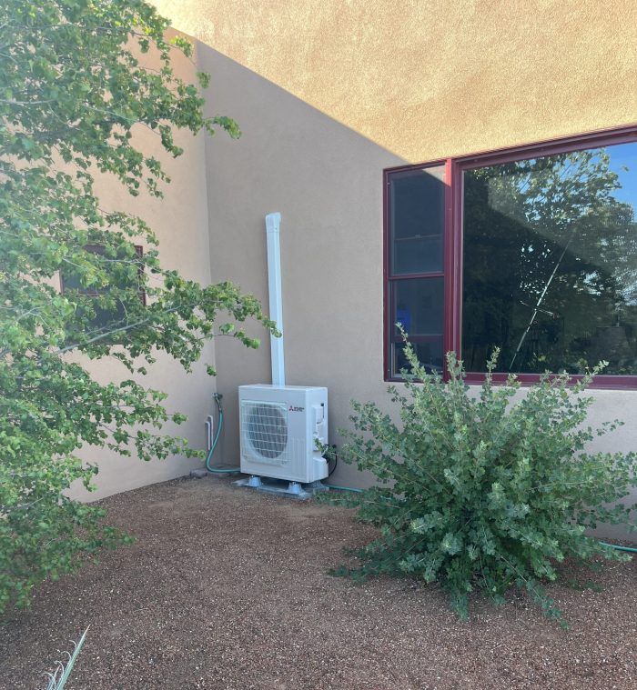 Air conditioning unit mounted on a beige wall next to a window, with white ducting.