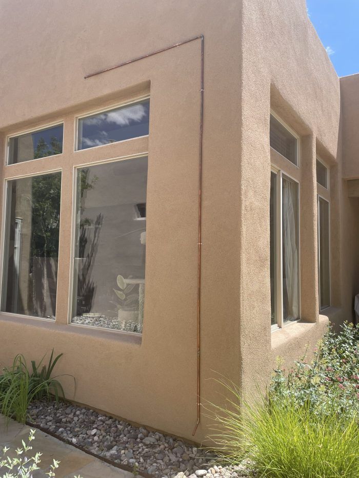 Tan stucco building exterior with windows, a copper pipe, and landscaping.