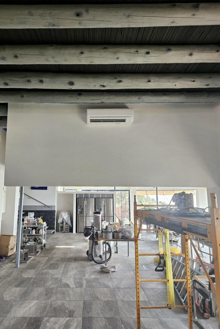 Interior view of a room with a white wall, AC unit, exposed wooden beams, and a view into another room.