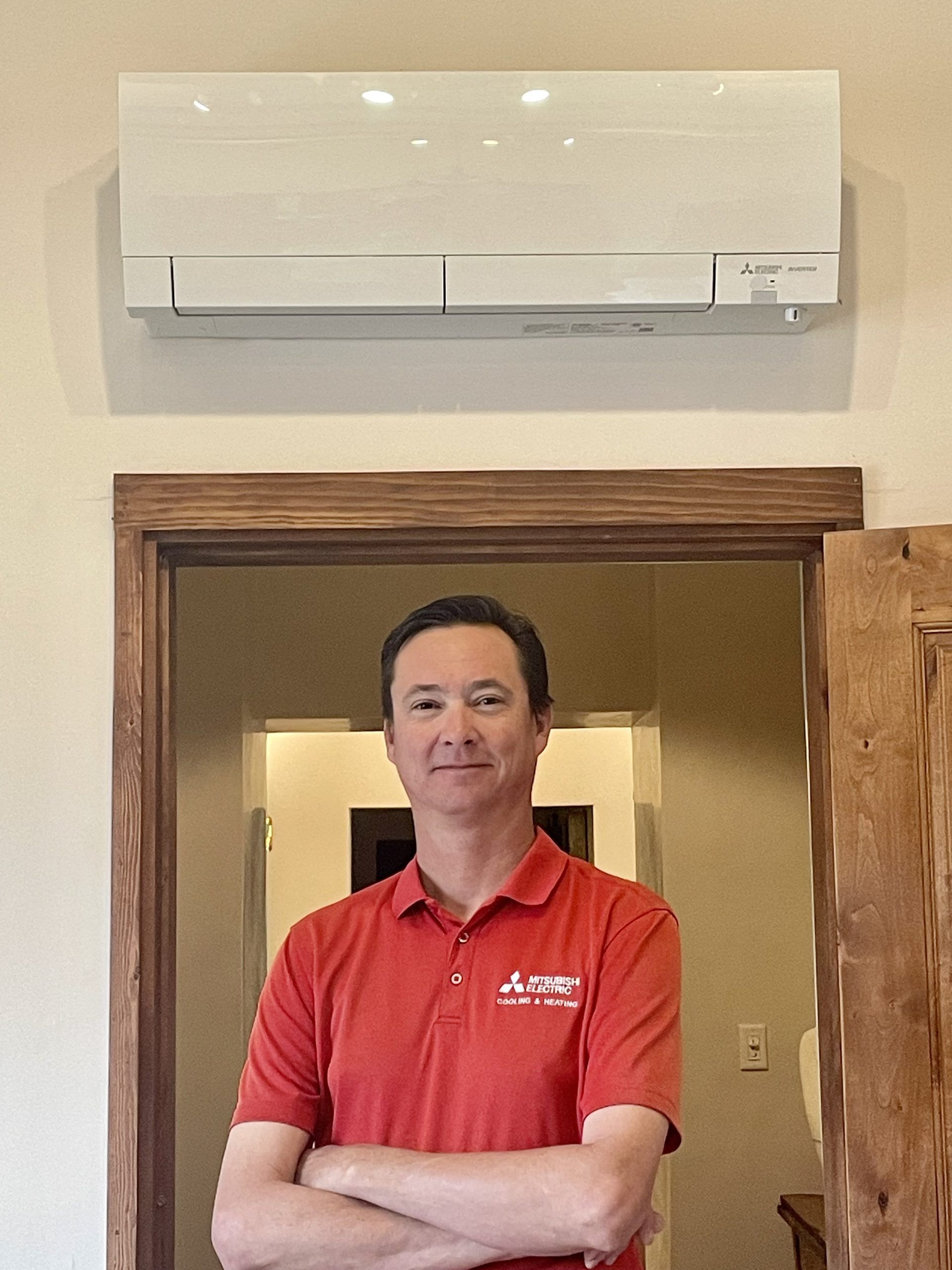 Man in red shirt stands with arms crossed under a wall-mounted Mitsubishi AC unit over a doorway.
