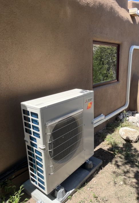 Exterior view of a beige heat pump unit next to a brown stucco wall, near a window, with a white conduit.