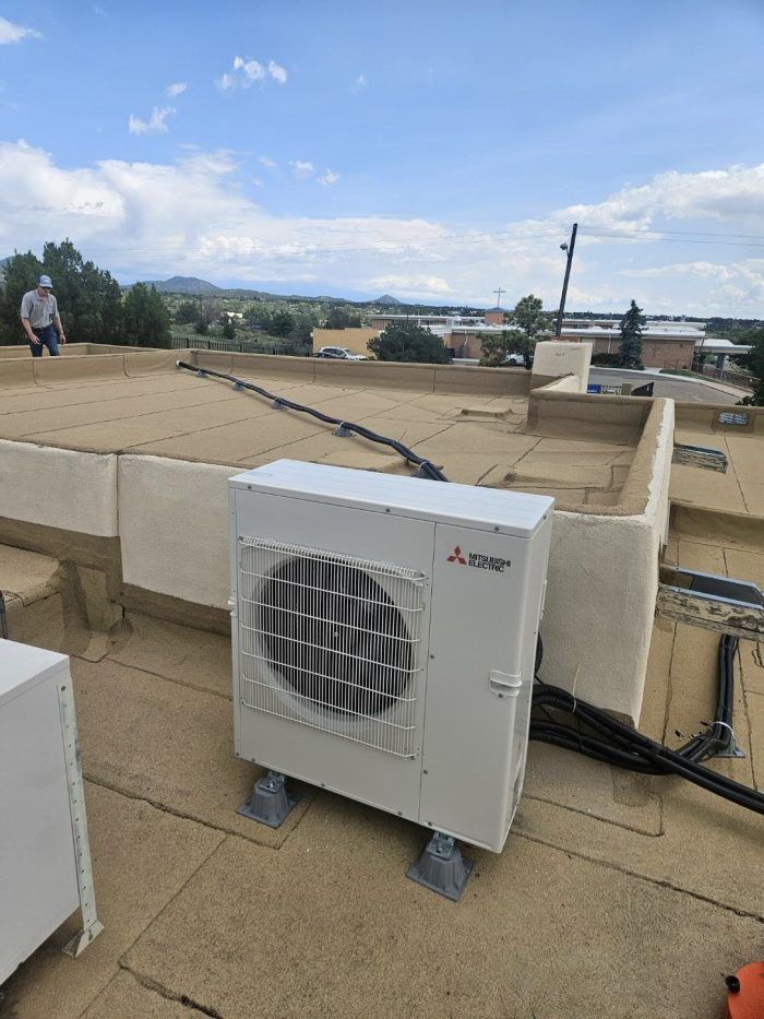 A Mitsubishi heat pump unit on a rooftop, with a worker in the background. Blue sky, wires, and tan surfaces.