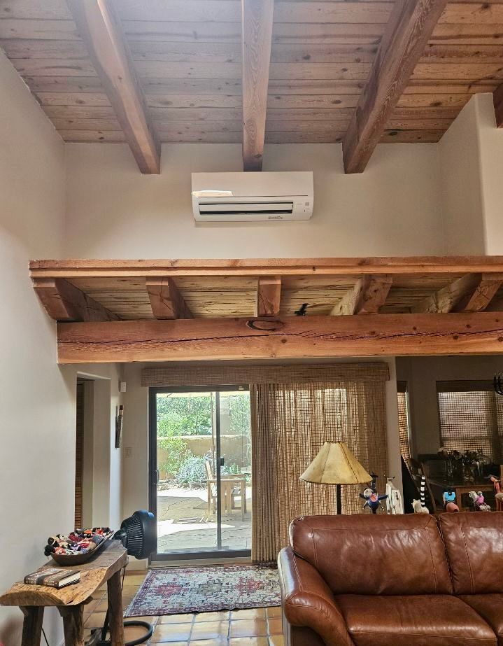 Air conditioner mounted on a wall above a wooden beam in a living room with a leather couch and a sliding glass door.