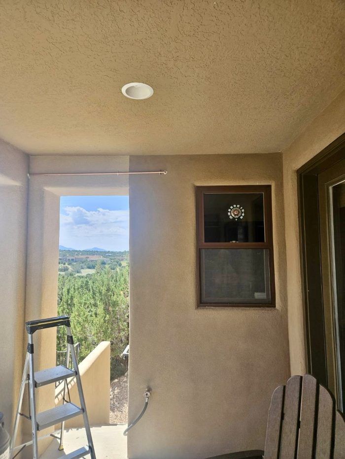 Beige stucco patio with a window, ladder, and a view of trees and sky.