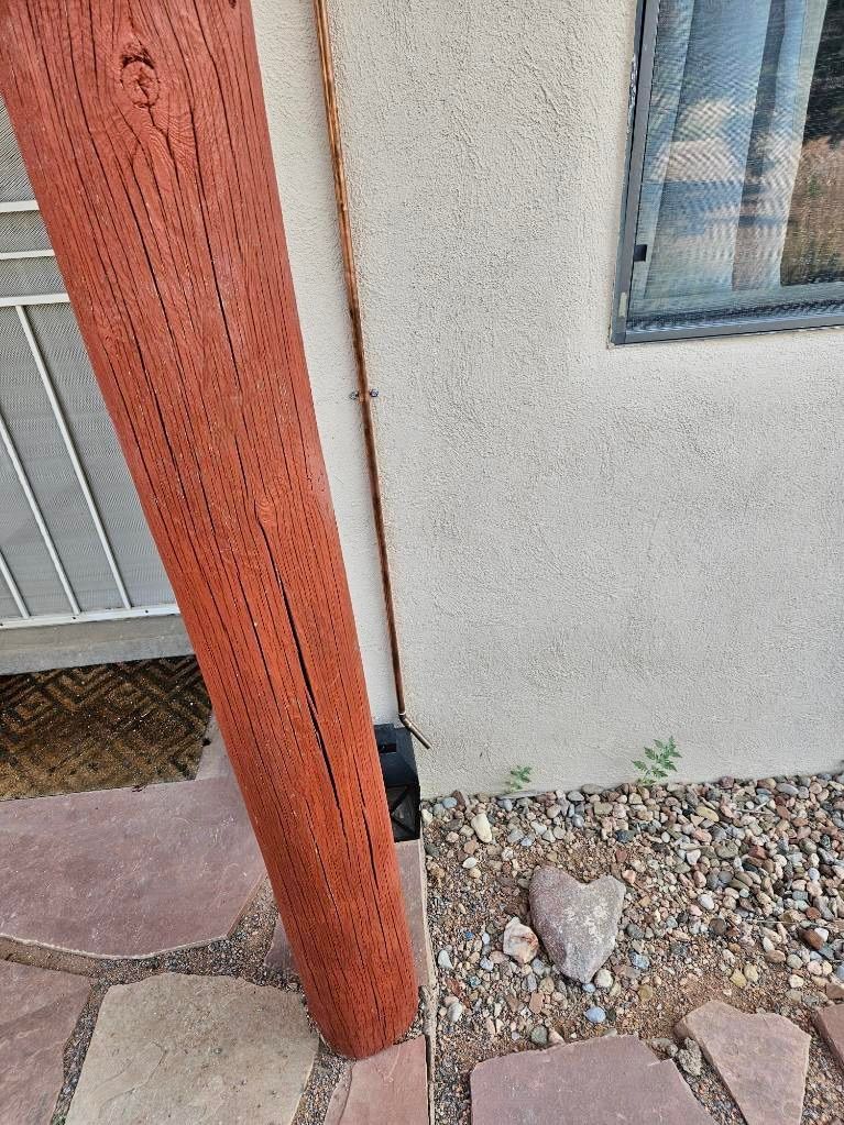 Red wooden post next to a stucco wall with a small window. Gravel and stone path below.