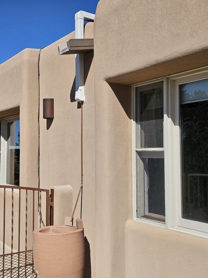 Tan stucco building with a white window frame, a rain barrel, and a downspout.