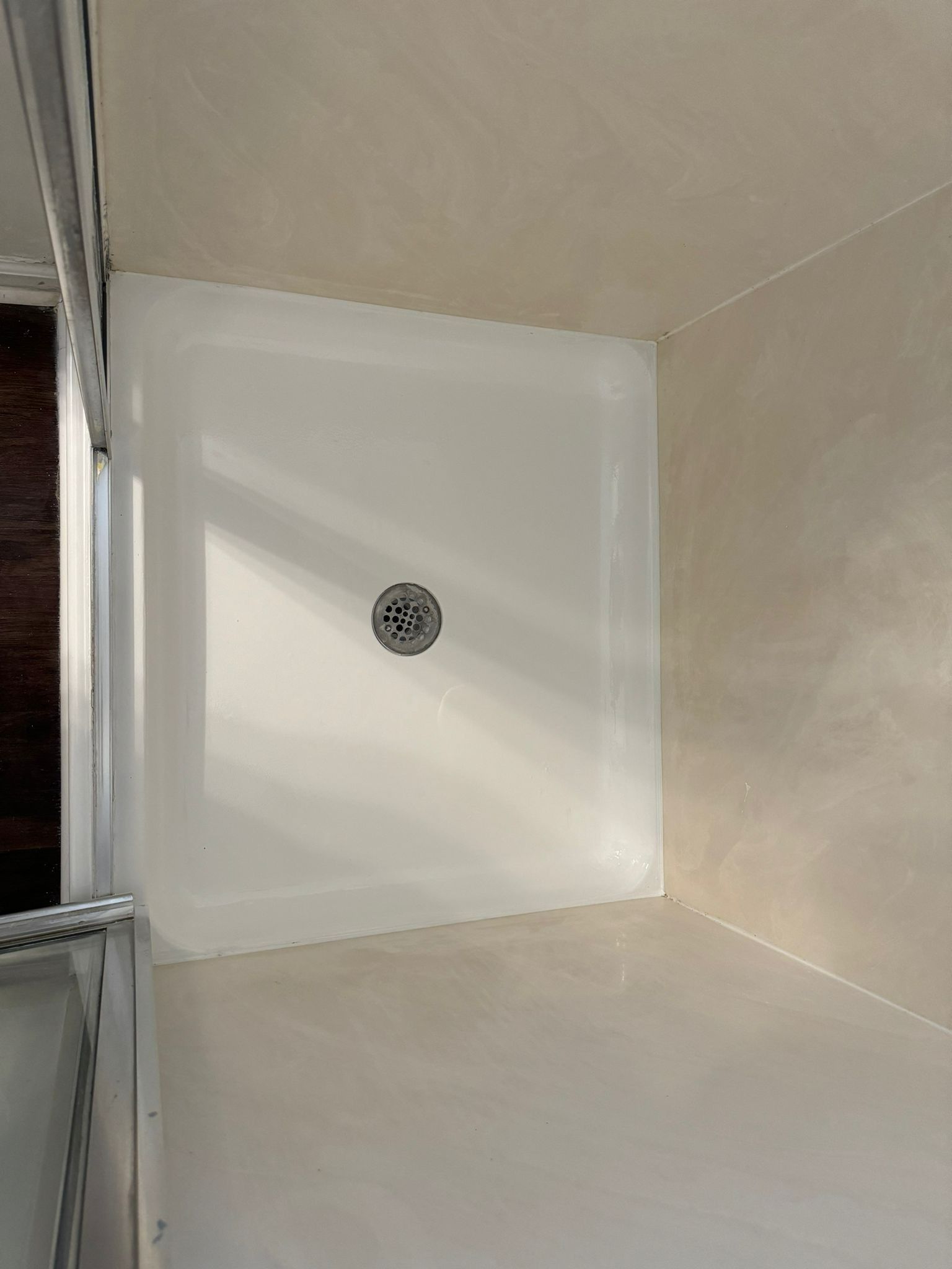 White shower base with drain, set in a tiled corner. Glass door frame on the left.