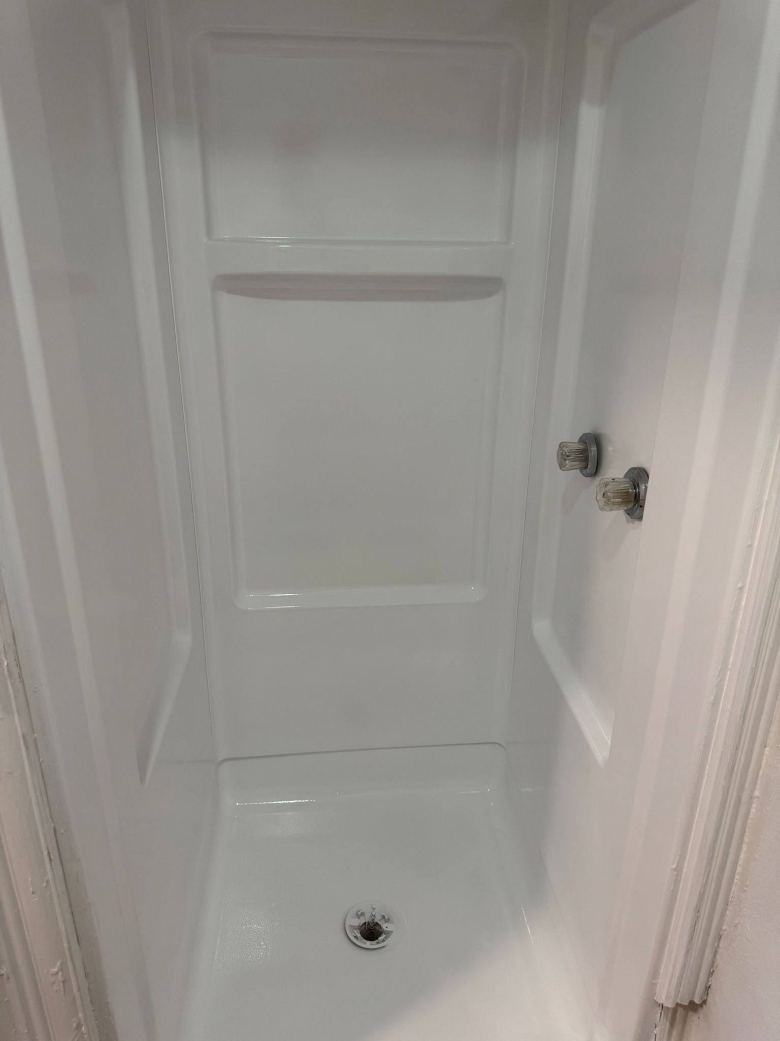 White, empty shower stall with built-in shelves, drain, and silver faucet.