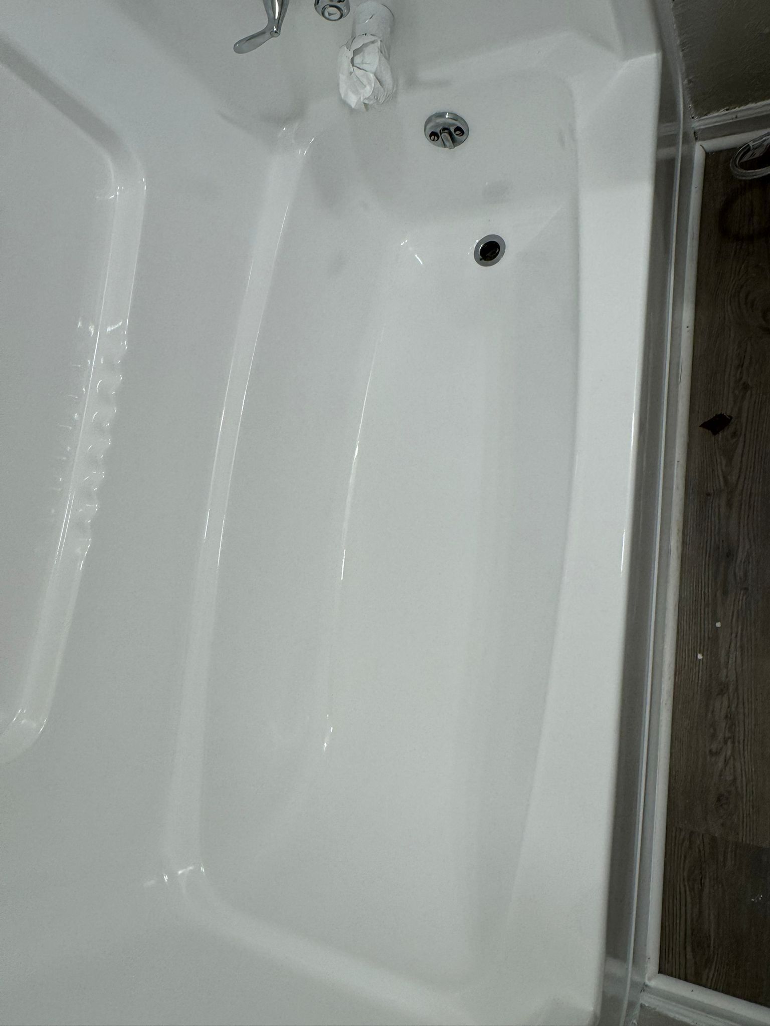White bathtub with showerhead and drain, in a bathroom with a glass door and wood-look flooring.