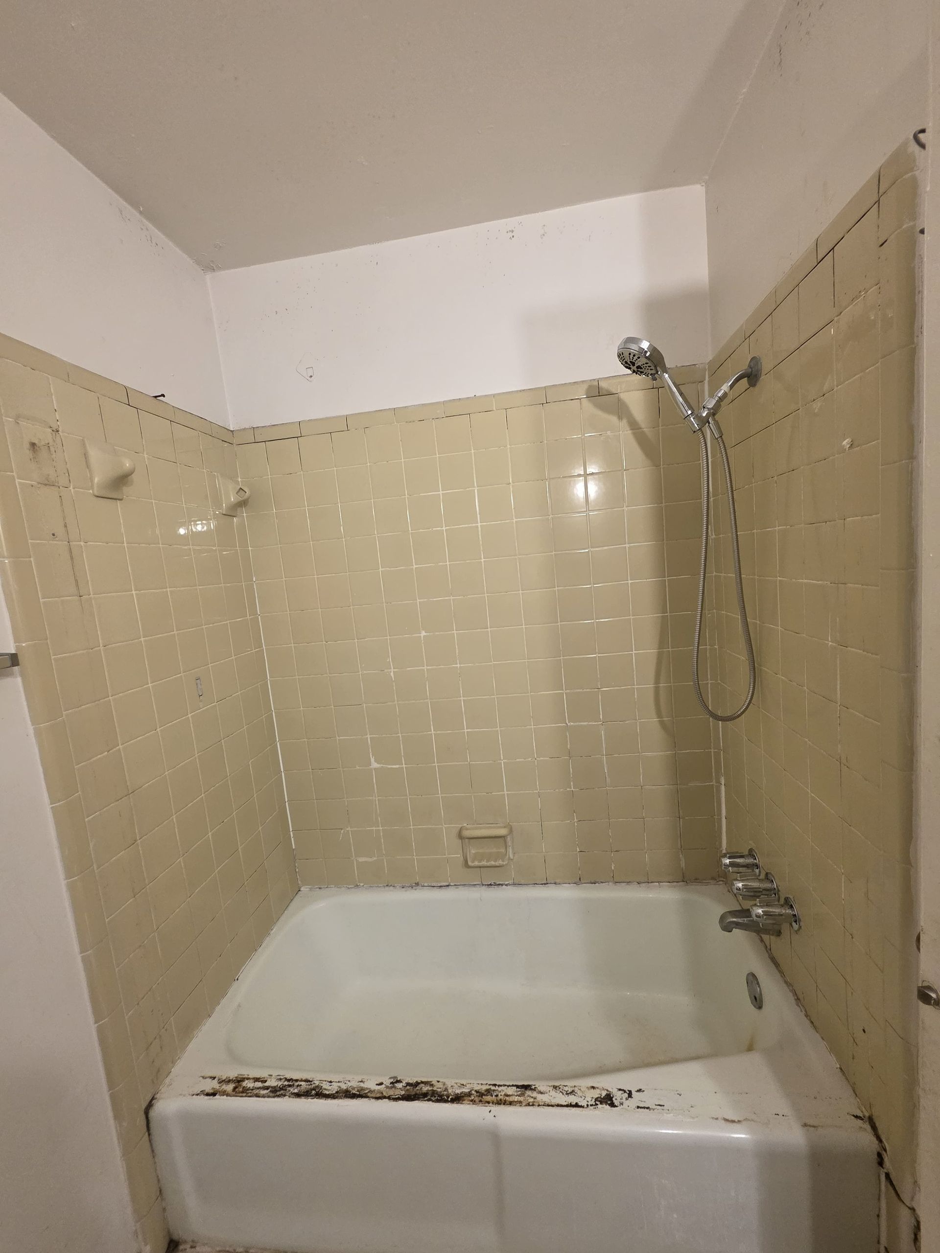 A dingy bathroom with a white bathtub, beige tiled walls, and showerhead.