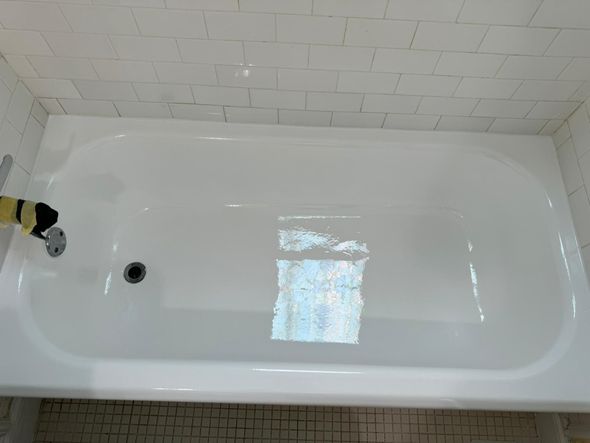 White bathtub in a bathroom, surrounded by white tile walls and a dark drain.