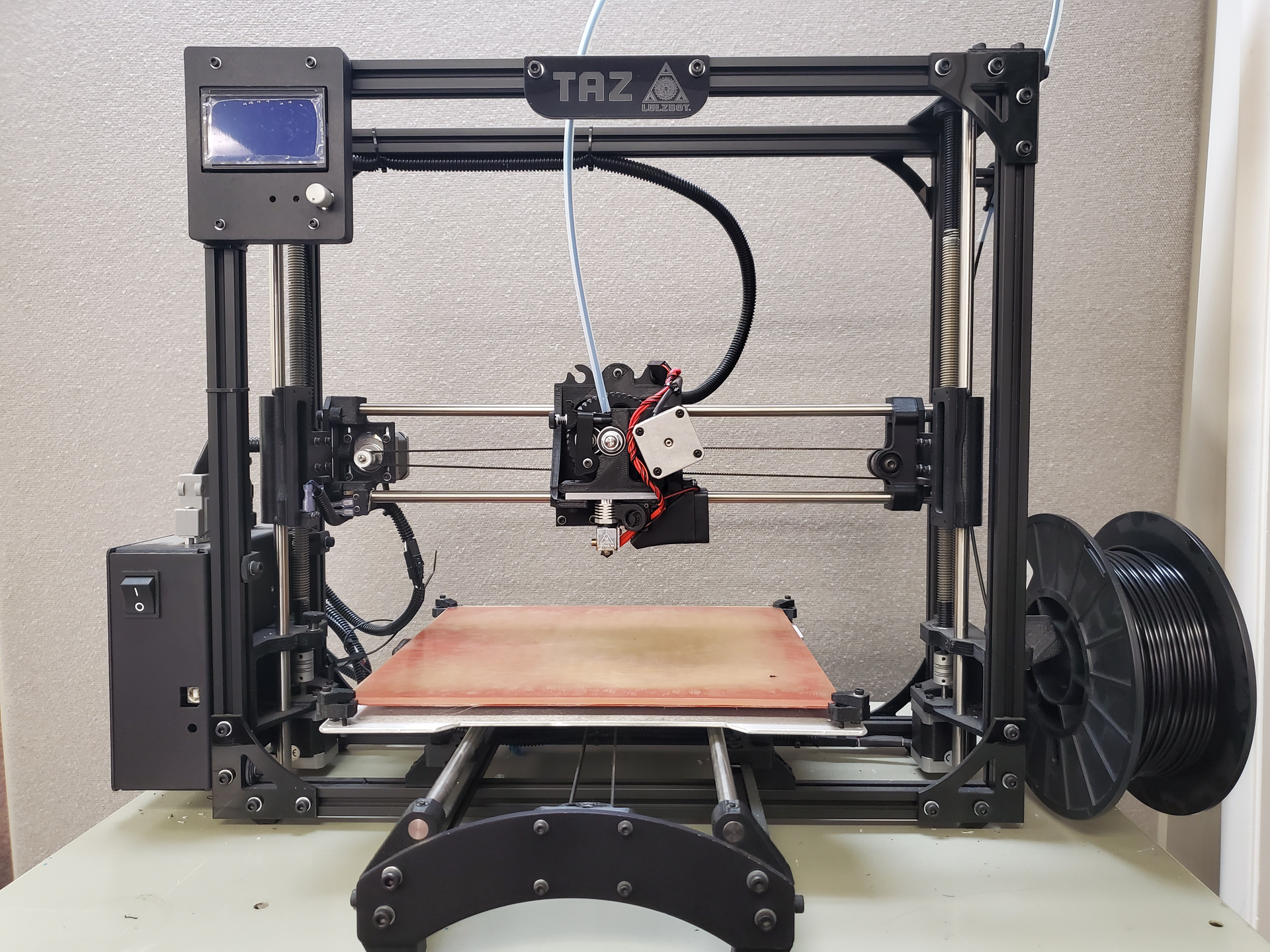 A black 3D printer, TAZ model, on a white table. A spool of black filament is on the right.