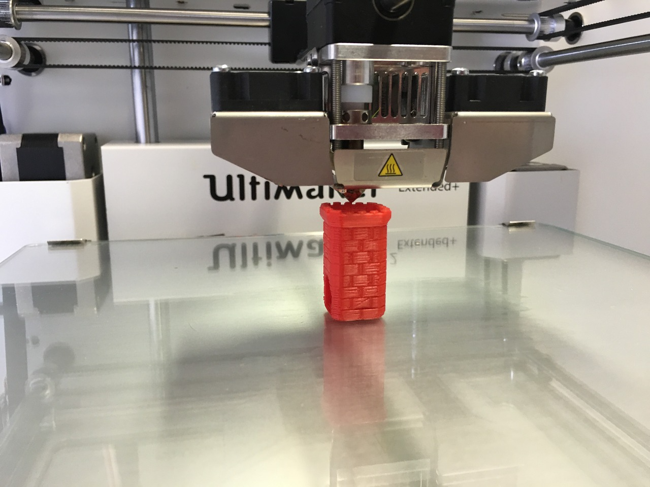 A 3D printer building a red plastic basket-like object on a shiny metal surface.