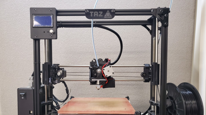 Black 3D printer, TAZ model, printing bed in center, spool of filament on right.