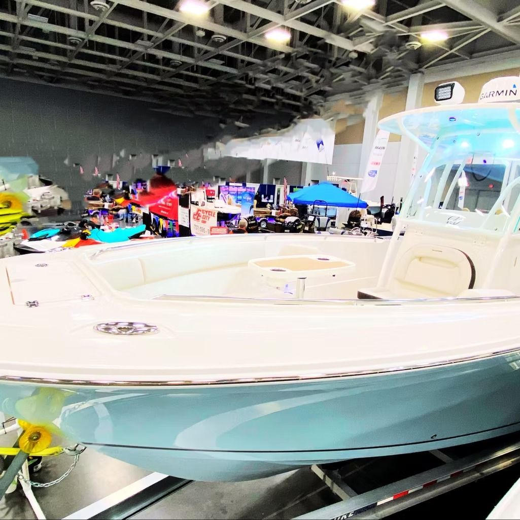 Boat at a boat show, light blue hull, white deck, people and booths in the background.
