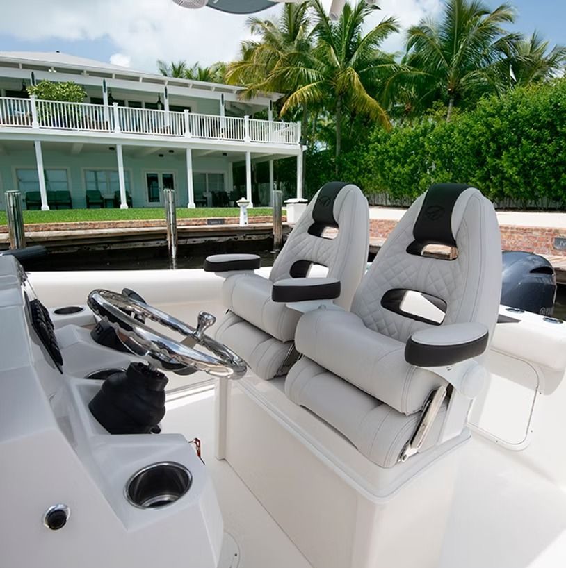 Boat helm with white seats, steering wheel, and a house in the background.