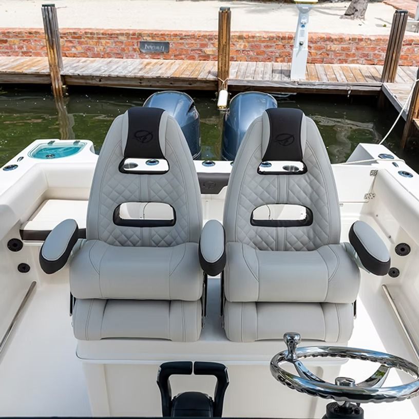 Two light gray boat seats with armrests and a steering wheel, docked near a brick wall.