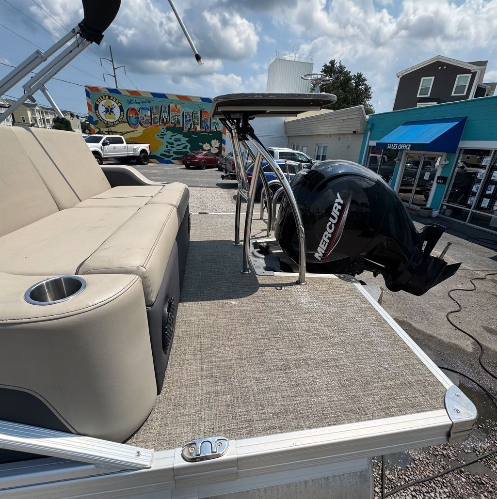 Tan pontoon boat with Mercury motor, parked outside businesses on a sunny day.