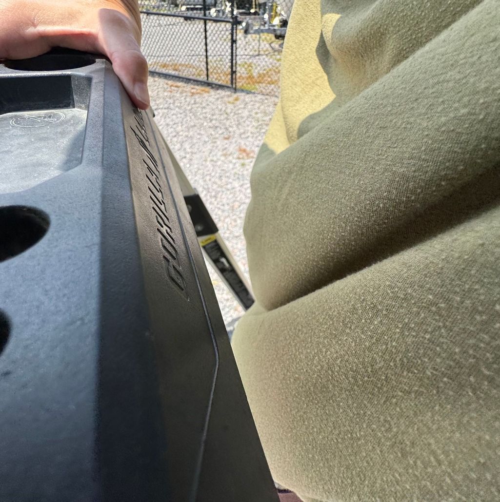 Person holding a black object with etched letters, wearing an olive green fleece jacket outdoors near a fence.