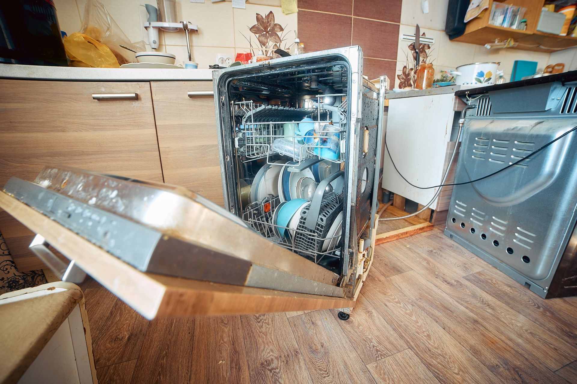 Broken dishwasher with open door in a kitchen, showing exposed interior parts and water damage.