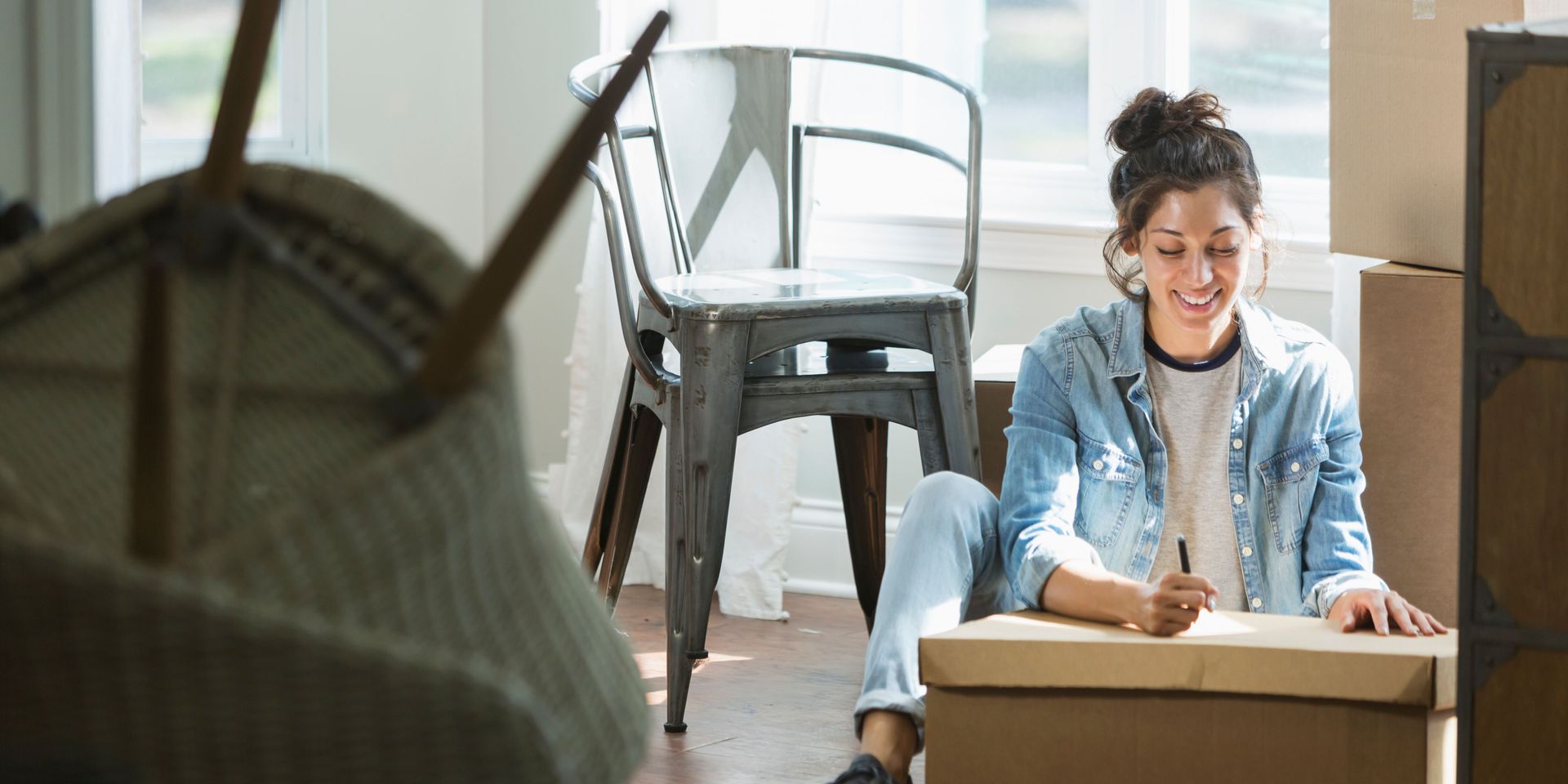 Woman sitting on the floor, writing on a moving box, smiling. Boxes and a chair are nearby.