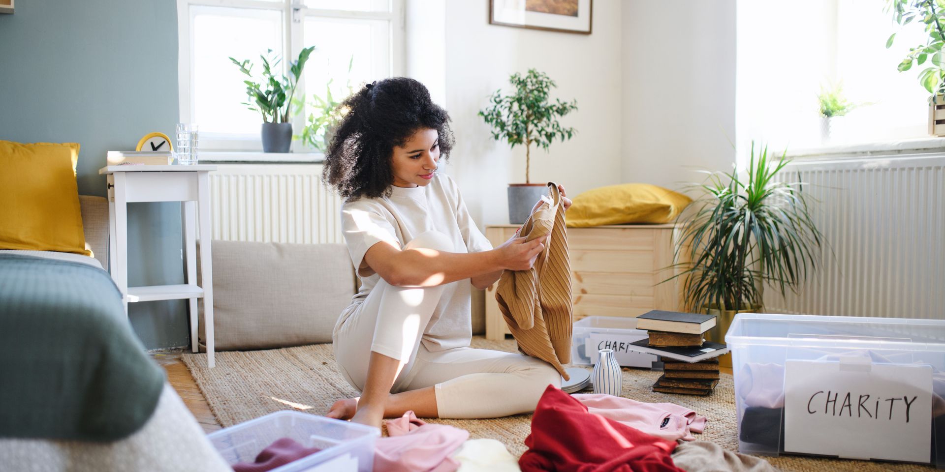 Woman sorting clothes for charity in her light-filled bedroom. She's looking at a tan sweater.