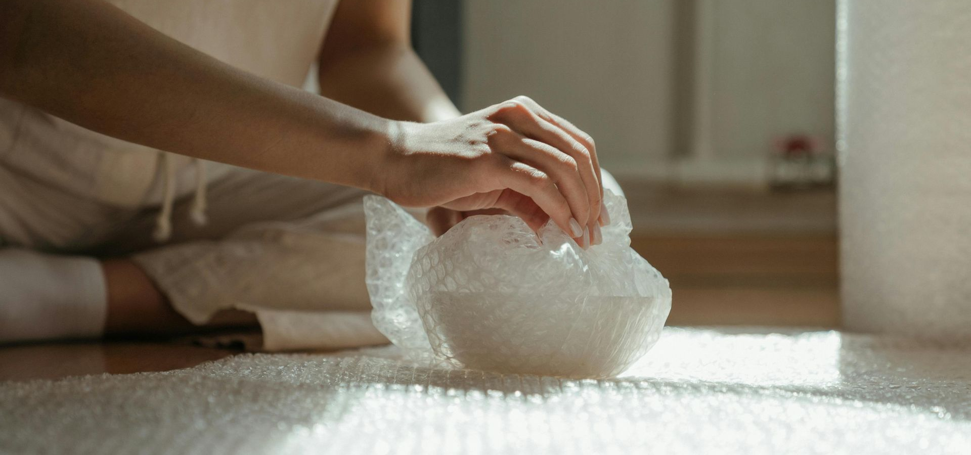 Person sitting on the floor wrapping an object in bubble wrap near a roll of bubble wrap.