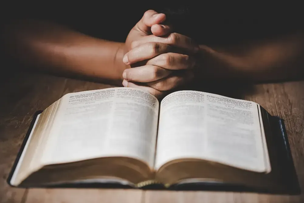 A person is praying over an open bible on a wooden table.