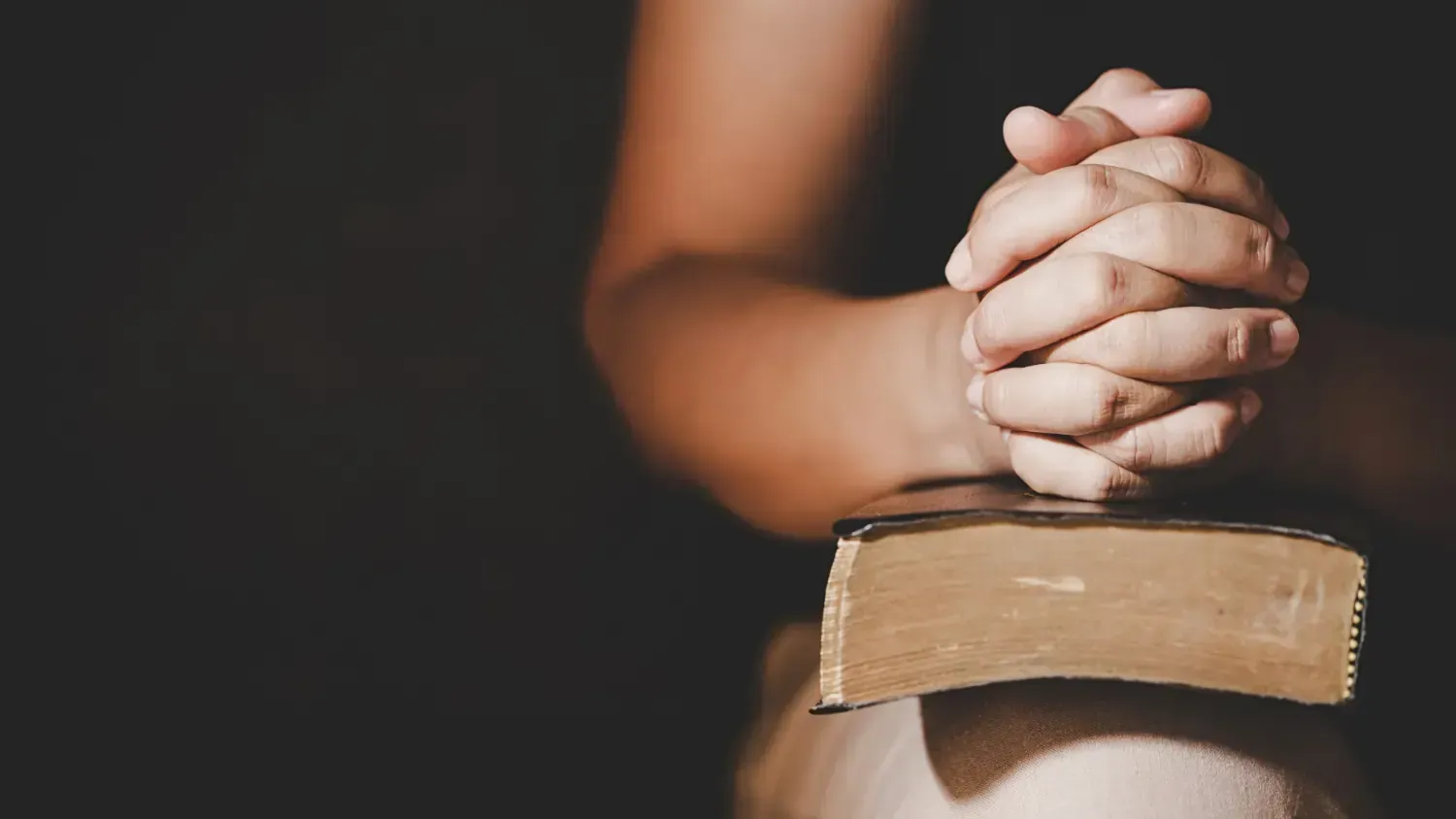 A person is praying with their hands folded over a bible.
