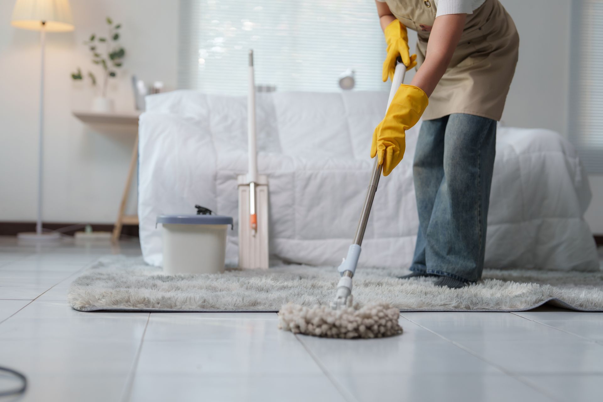 Person mopping a gray rug in a bright bedroom with a white bed and bucket nearby