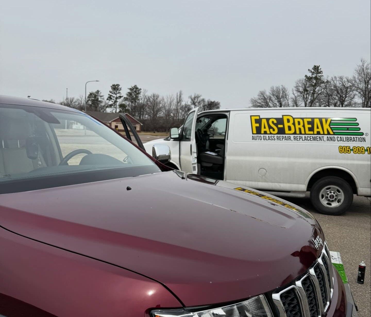 A burgundy Jeep parked next to a white Fas-Break glass repair van with its driver-side door open in a parking lot.