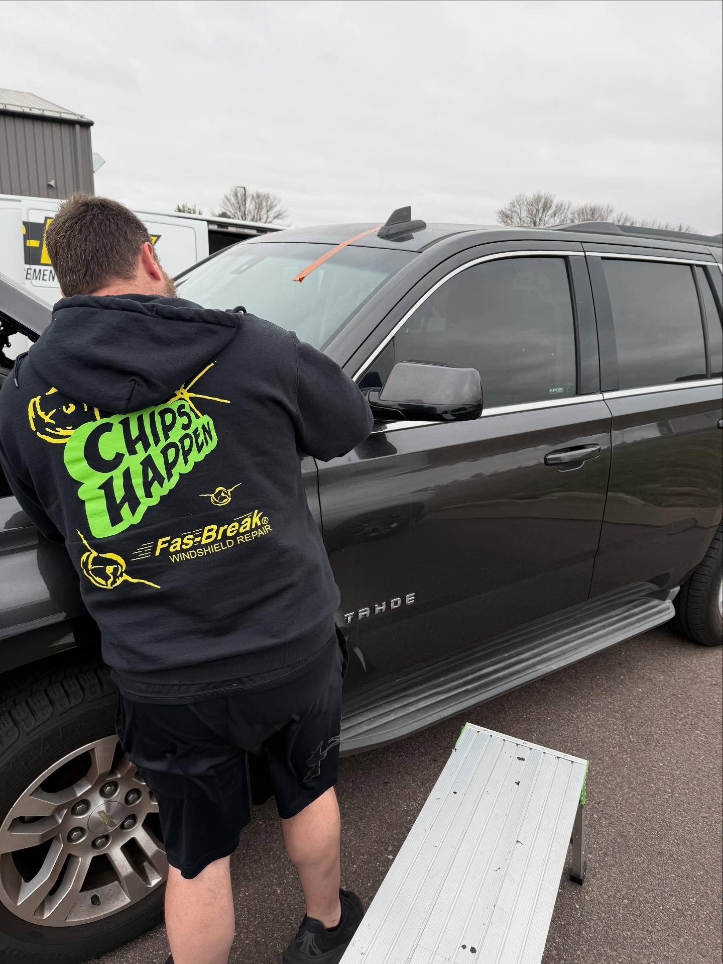 A technician in a branded black hoodie works on the windshield of a black SUV parked outdoors near a metal step platform.