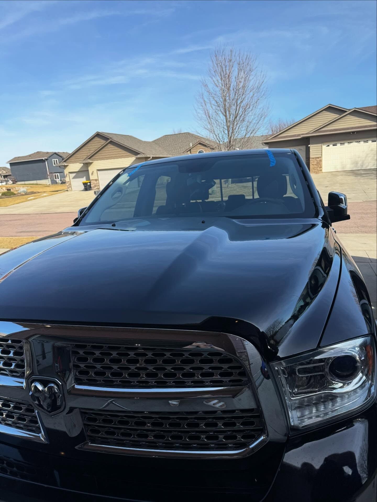 A close-up of a black Ram pickup truck parked in a suburban neighborhood driveway under a clear blue sky.