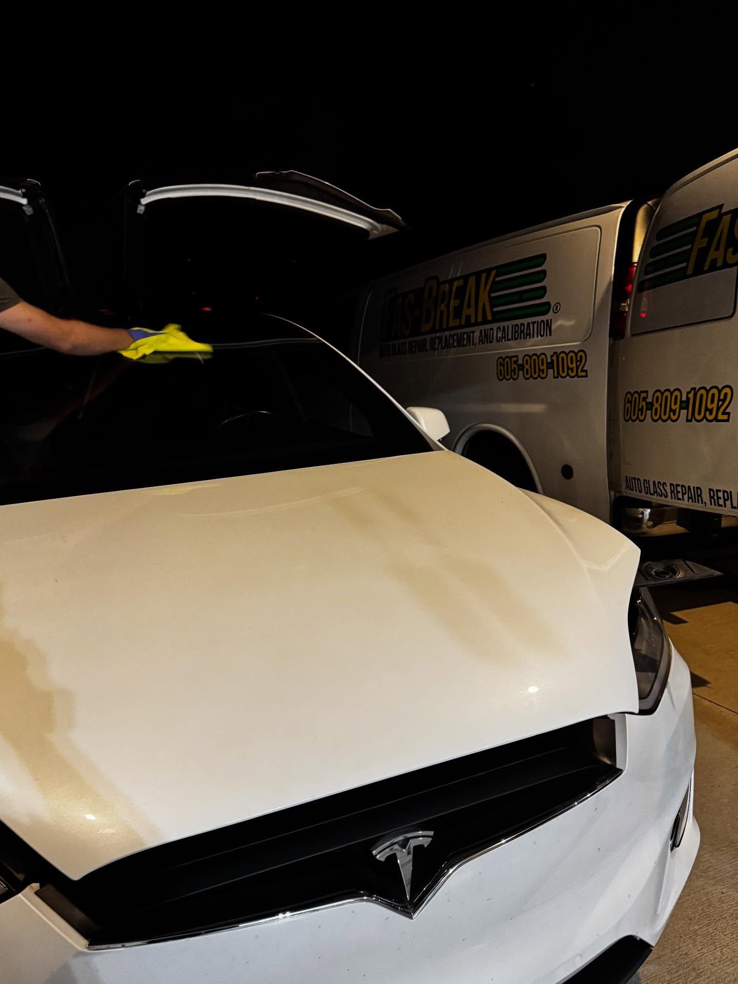 A person using a yellow cloth to wipe the windshield of a white Tesla parked at night next to a work van.