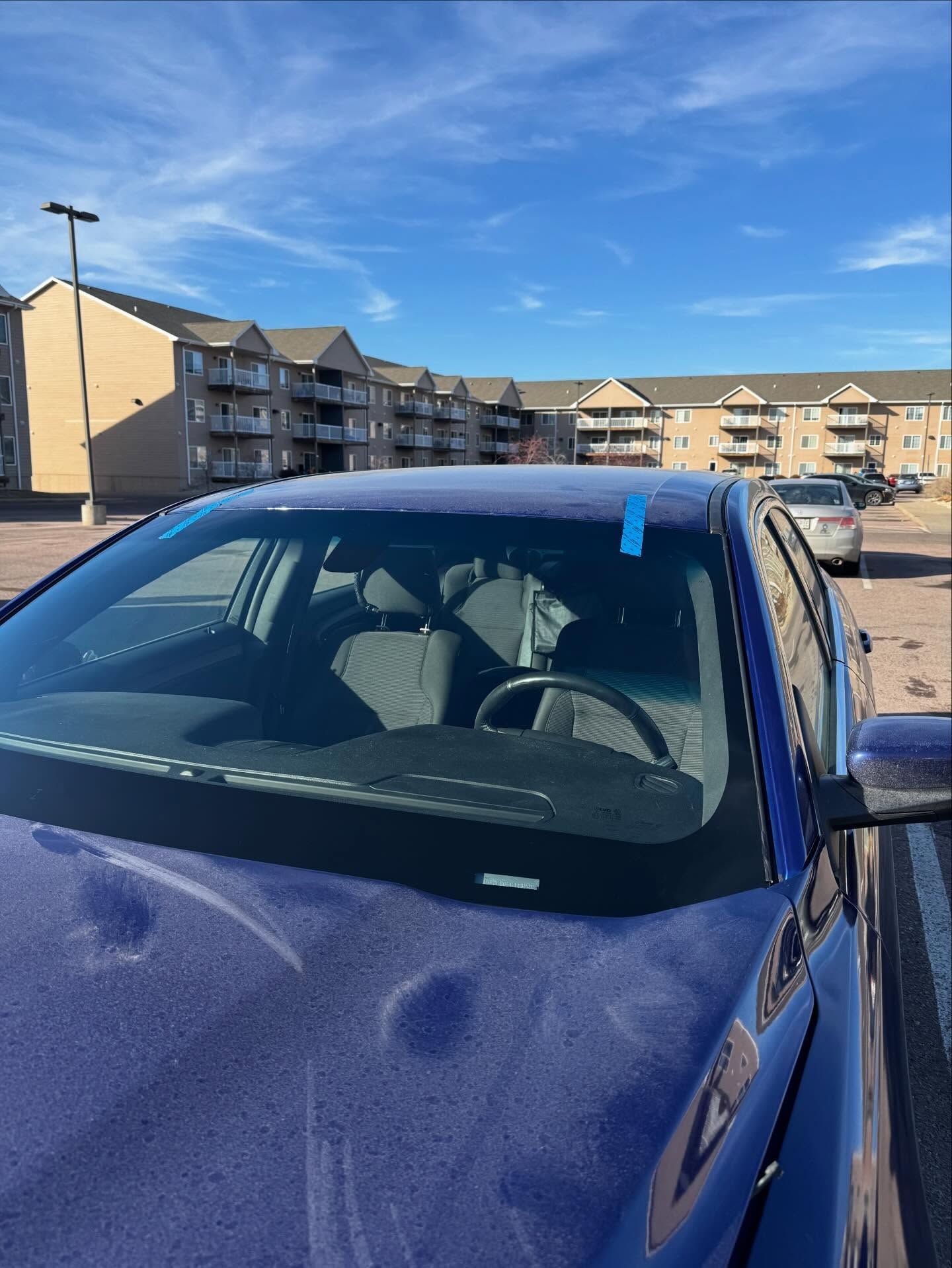 A dark blue car with visible hail dents on the hood, parked in front of apartment buildings under a clear blue sky.