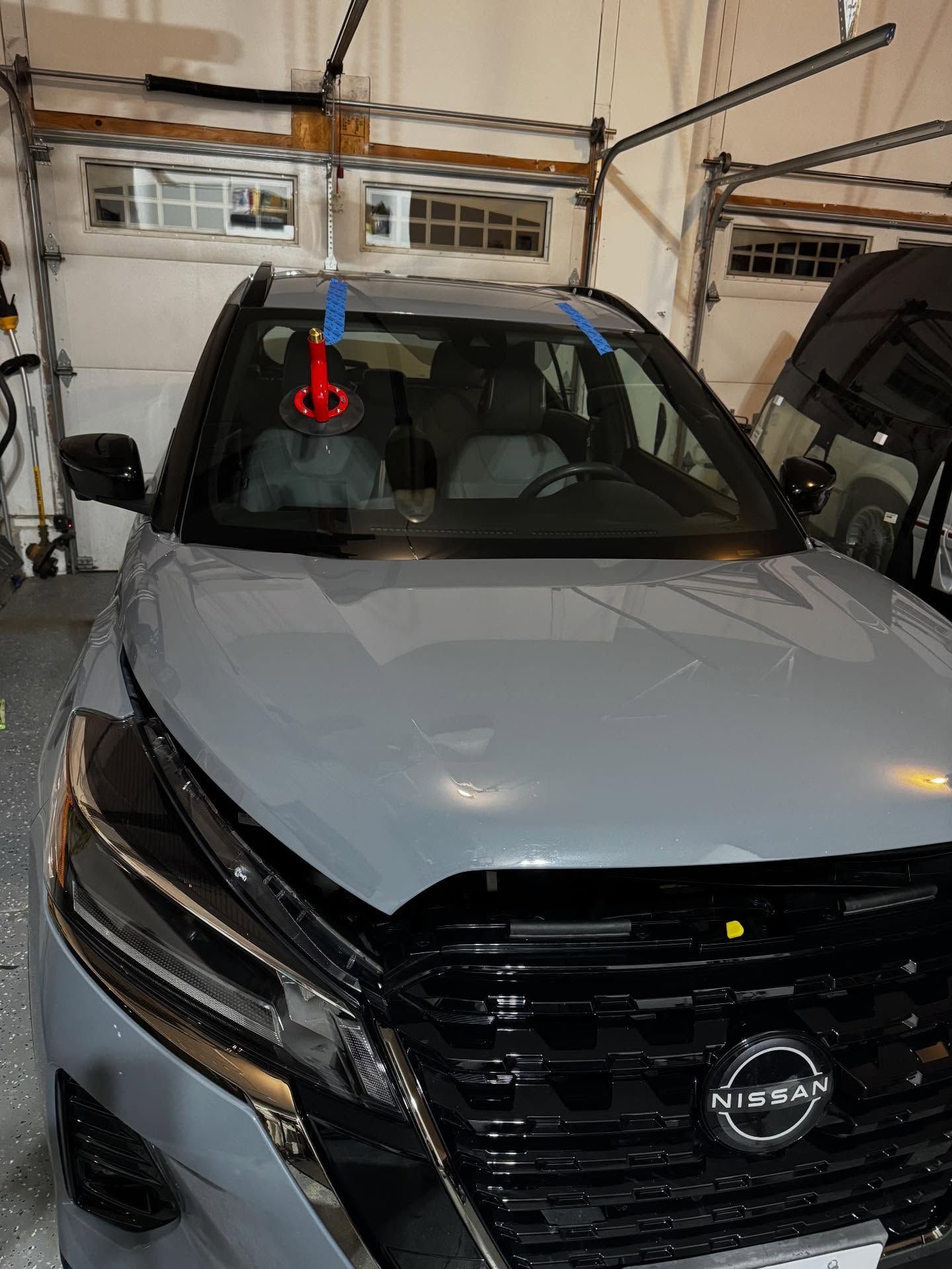 A gray Nissan SUV parked in a garage with a red suction cup tool attached to the windshield for glass repair.