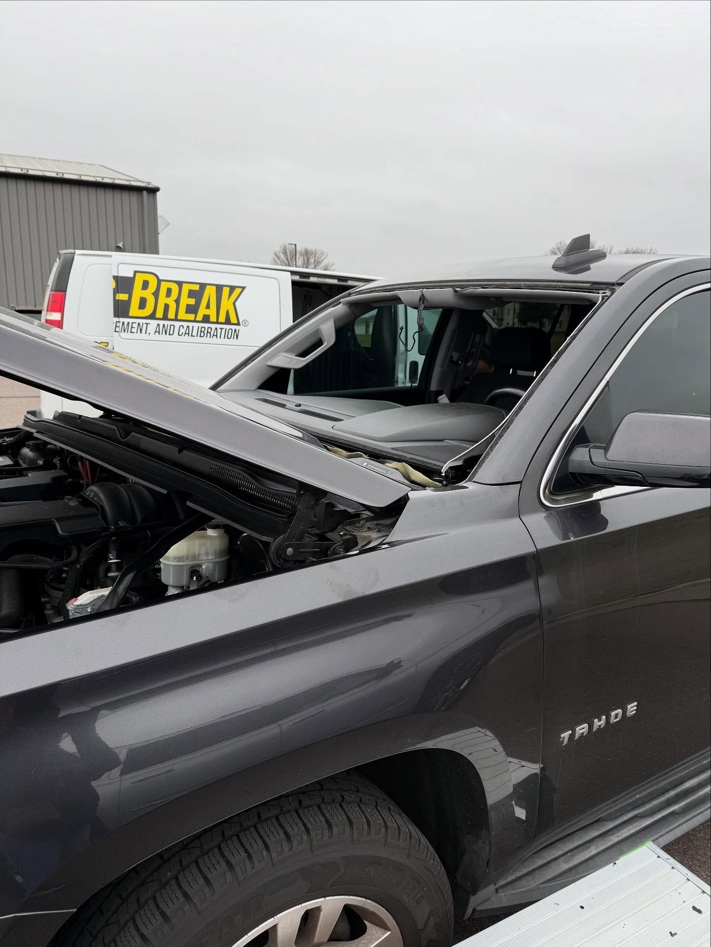 A dark gray SUV with its hood popped and front windshield completely shattered, parked near a service van.