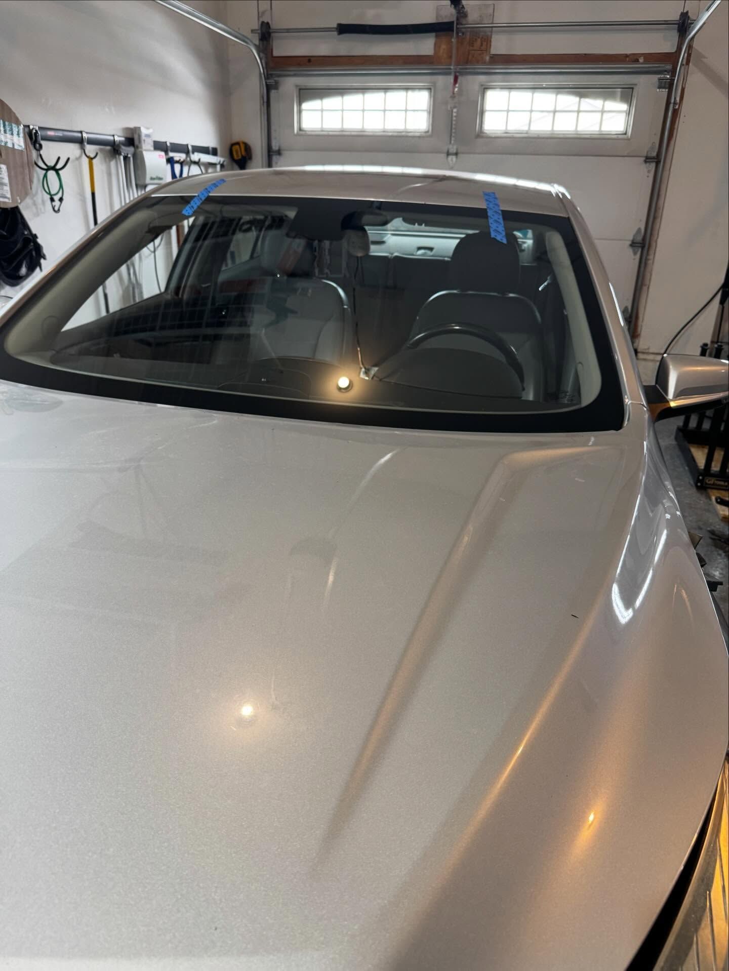 A silver car parked inside a garage, with blue painter's tape marking the top of the windshield.
