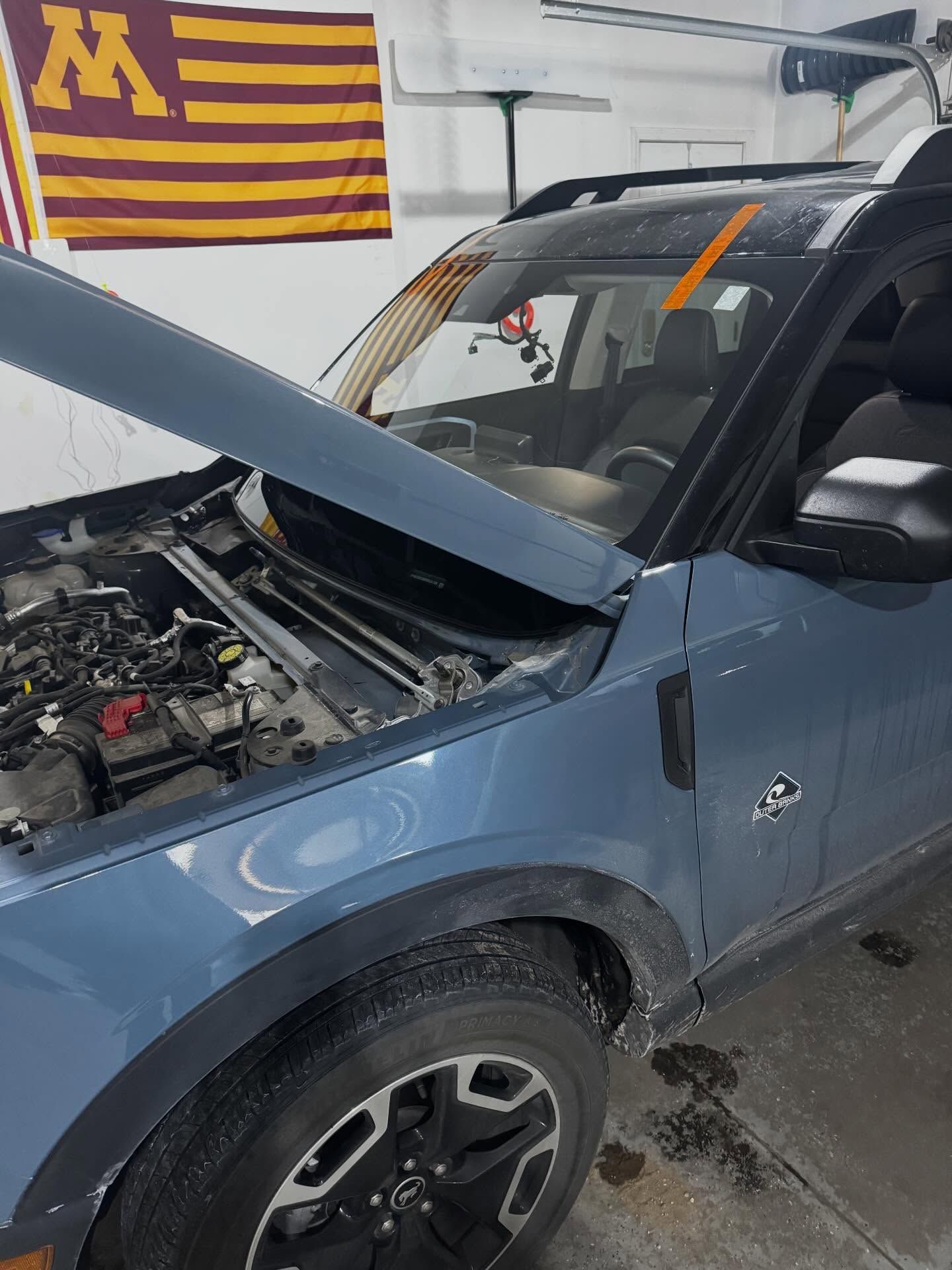 A blue Ford Bronco Sport with its hood open, parked in a garage with a University of Minnesota flag on the wall.