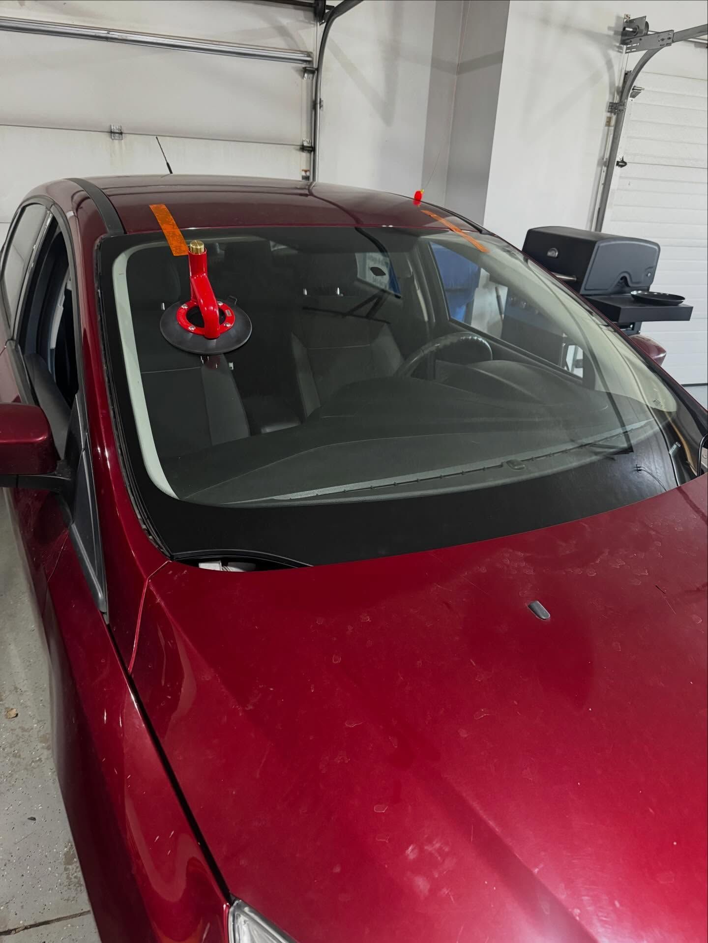A red car inside a garage with a red suction cup tool attached to its windshield, indicating a glass repair in progress.