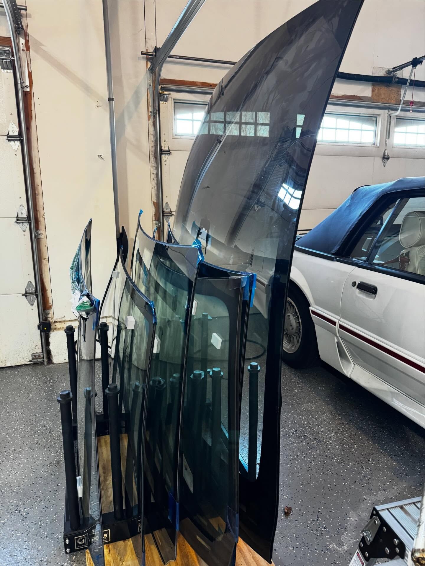 A rack holding several large, glass automotive windshields stands in a garage beside a white convertible car.
