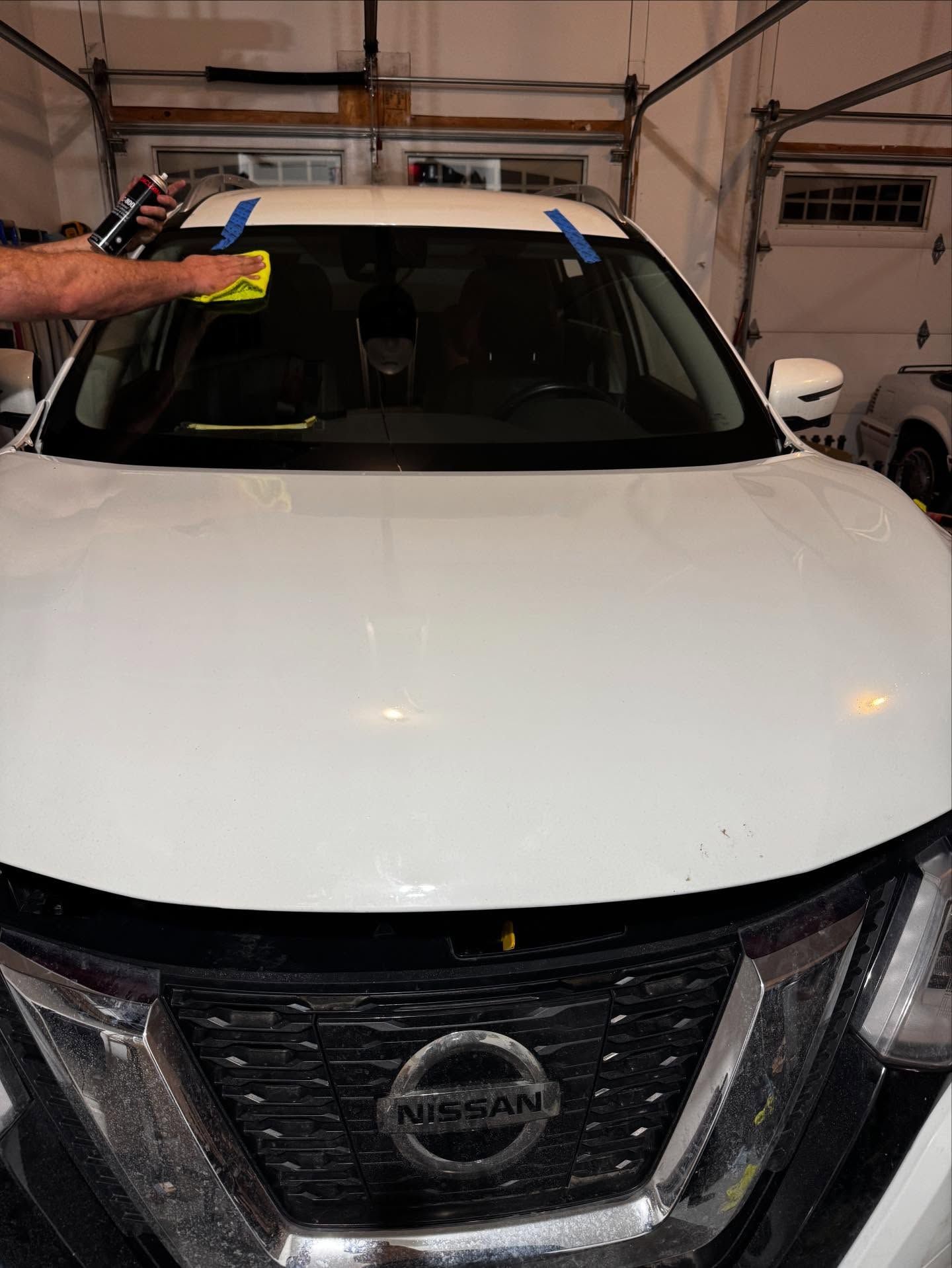 A hand wipes the top of a white car windshield with a yellow cloth in a garage.